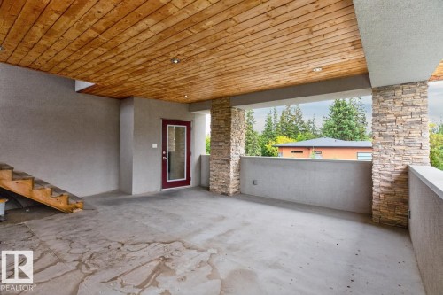 Covered outdoor space featuring a wood-panel ceiling with recessed lighting, stone-clad pillars, a red-framed glass door, and exposed wood stairs - 14A Birch Drive, St. Albert, AB 