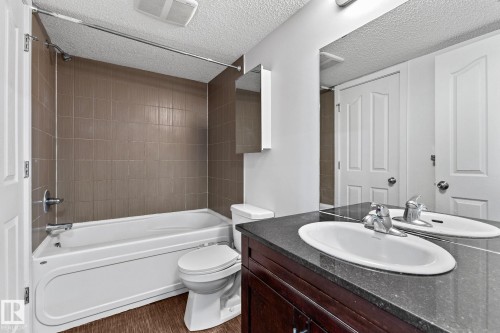 Tub/shower combination with tile surround, wood-finish flooring, dark wood vanity with white oval sink, and large wall-mounted mirror - #306 530 Watt Bv Sw, Edmonton, AB 