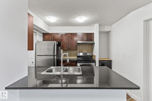 Kitchen featuring dark wood-finish cabinetry, stainless steel appliances, a double basin sink, and dark countertops - #306 530 Watt Bv Sw, Edmonton, AB 