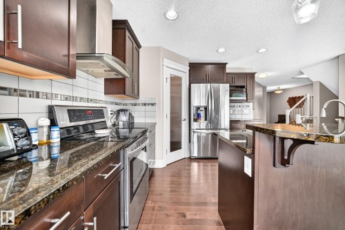 Kitchen featuring stainless steel appliances, dark wood-finish cabinetry, granite countertops, tile backsplash with decorative accent, and wood-finish flooring - 12035 168 Avenue Nw, Edmonton, AB 