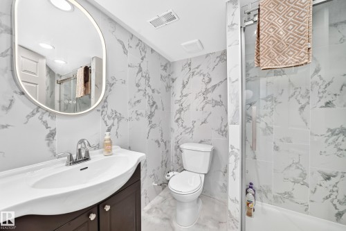Bathroom featuring floor-to-ceiling marbled-patterned tile, a dark wood-finish vanity with a white countertop and integrated sink, a frameless oval mirror with integrated lighting, and a glass-enclosed shower with a tub - 12035 168 Avenue Nw, Edmonton, AB 