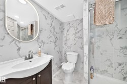 Bathroom featuring floor-to-ceiling marbled-patterned tile, a dark wood-finish vanity with a white countertop and integrated sink, a frameless oval mirror with integrated lighting, and a glass-enclosed shower with a tub - 