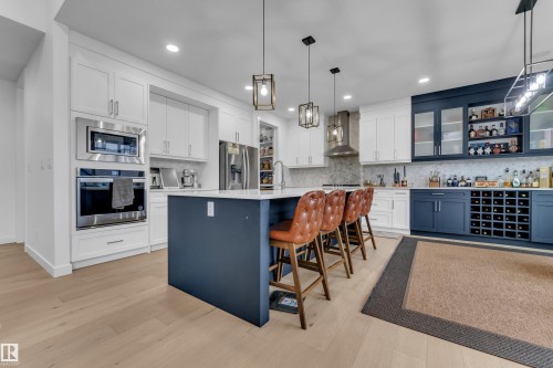 Modern kitchen featuring light wood-finish flooring, white cabinetry, a built-in oven and microwave, a large central island with a contrasting base, and a dedicated beverage station with blue cabinetry and a wine rack - 1409 Graydon Hill Way Sw, Edmonton, AB 