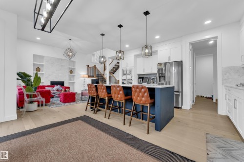 Open-concept living area with wood-finish flooring, a white brick-style fireplace with built-in shelving, a kitchen island with pendant lighting, white cabinetry, and stainless steel appliances - 1409 Graydon Hill Way Sw, Edmonton, AB 
