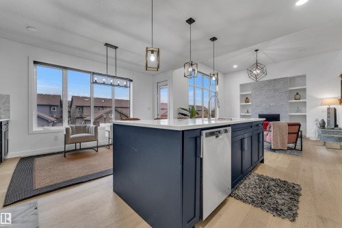 Open-concept kitchen and living area featuring a large center island with dark cabinetry and white countertops, pendant lighting, wood-finish flooring, and abundant natural light from expansive windows overlooking the neighborhood - 1409 Graydon Hill Way Sw, Edmonton, AB 