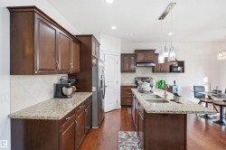 Kitchen featuring wood-finish flooring, dark wood cabinetry, light-toned countertops, a stainless steel refrigerator, and a kitchen island with an integrated sink - 