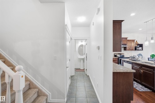 Carpeted staircase with white railings, tile flooring in the hallway, pedestal sink with oval mirror, and a kitchen featuring dark wood cabinetry, a granite-style countertop, and pendant lighting - 1523 36 Avenue Nw, Edmonton, AB 