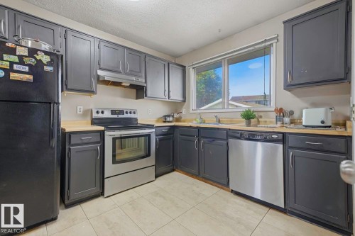 Kitchen featuring dark cabinetry, light-toned countertops, tile flooring, and stainless steel appliances including a range, dishwasher, and refrigerator - 3216 138 Avenue Nw, Edmonton, AB 
