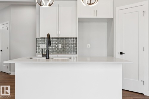 Kitchen island featuring a single-basin sink with a matte black faucet, white cabinetry, and a textured tile backsplash - 72 Maple Crescent, Gibbons, AB 