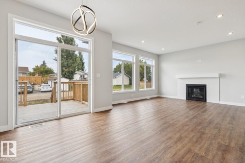 Spacious room featuring wood-finish flooring, a contemporary fireplace with a white mantel, multiple large windows, and a sliding glass door leading to a wooden deck - 72 Maple Crescent, Gibbons, AB 