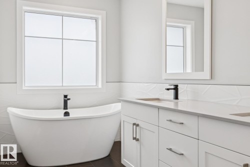 Freestanding soaking tub with a matte black faucet, double vanity with integrated sinks, white quartz countertop, and textured tile wainscoting - 72 Maple Crescent, Gibbons, AB 