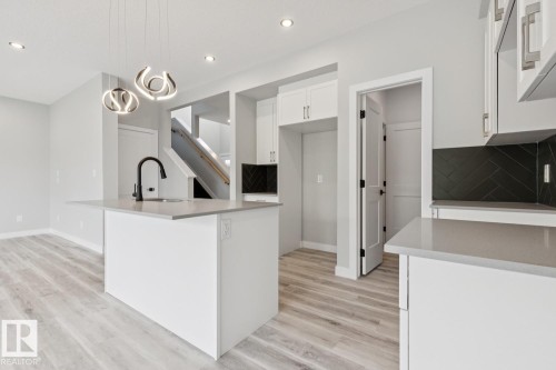 Kitchen featuring a central island with a sink, light-toned countertops, white cabinetry, a black herringbone tile backsplash, and wood-finish flooring - 36 Maple Crescent, Gibbons, AB 