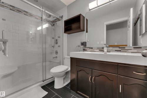 Double vanity with integrated sinks, glass-enclosed shower, multi-function shower panel, contemporary wall-mounted light fixture, and dark wood-finish cabinetry - 9722 157 Street Nw, Edmonton, AB 