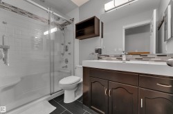Double vanity with integrated sinks, glass-enclosed shower, multi-function shower panel, contemporary wall-mounted light fixture, and dark wood-finish cabinetry - 