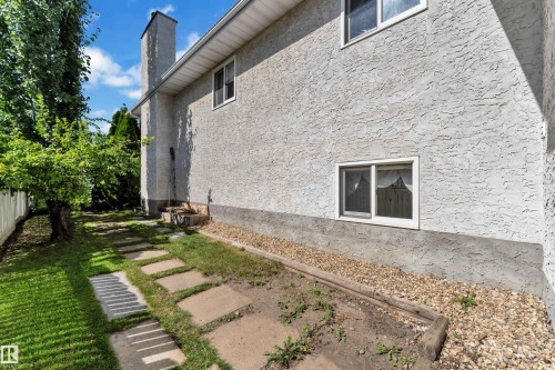 Exterior featuring stucco finish, white frame windows, rain gutters, a prominent chimney, and a landscaped side yard with stepping stones and mature trees - 9722 157 Street Nw, Edmonton, AB 