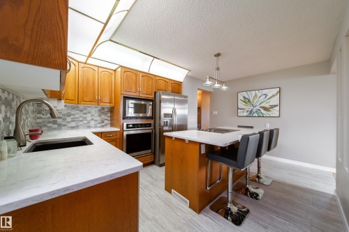 Wood cabinetry, light-toned countertops, stainless steel appliances, mosaic tile backsplash, and light wood-finish flooring - 10438 32A Avenue Nw, Edmonton, AB 