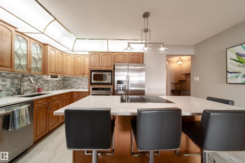 Kitchen featuring wood cabinetry, stainless steel appliances, a multi-tone tile backsplash, and a large island with an integrated cooktop - 10438 32A Avenue Nw, Edmonton, AB 