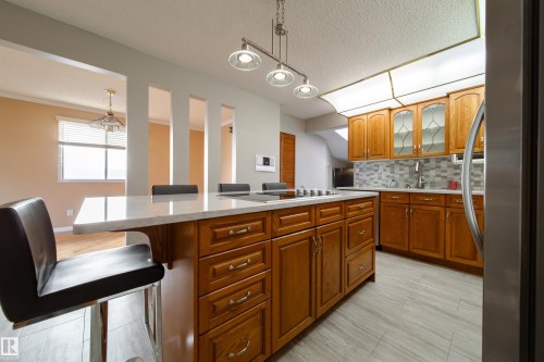 Kitchen featuring wood cabinetry, a large center island with a light-toned countertop, a tile backsplash, stainless steel appliances, and tile flooring - 10438 32A Avenue Nw, Edmonton, AB 