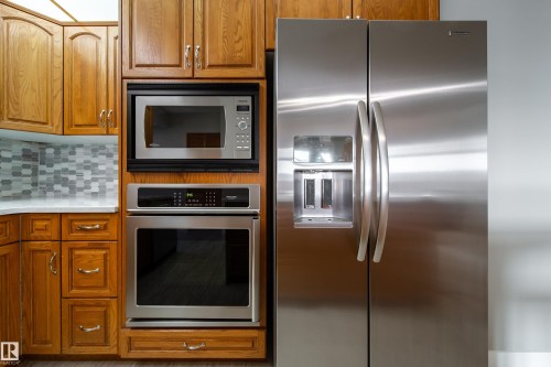 Stainless steel side-by-side refrigerator with water and ice dispenser, built-in microwave, wall oven, wood cabinetry, and mosaic tile backsplash - 10438 32A Avenue Nw, Edmonton, AB 