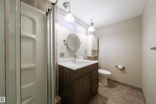 Bathroom featuring a shower enclosure, wood-finish vanity with white countertop, round wall mirror, and tiled flooring - 10438 32A Avenue Nw, Edmonton, AB 