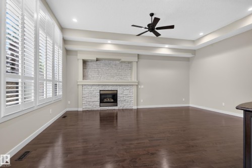 Hardwood flooring, a stone-clad fireplace with mantel, white louvered window treatments, a tray ceiling with recessed lighting, and a ceiling fan - 904 Armitage Court Sw, Edmonton, AB 