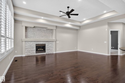 Stacked stone fireplace with mantel, tray ceiling with recessed lighting, dark wood-finish flooring, ceiling fan, and plantation shutters - 904 Armitage Court Sw, Edmonton, AB 