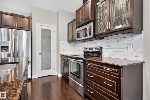 Kitchen featuring dark wood-finish cabinetry, stainless steel appliances, patterned glass upper cabinet doors, granite countertops, and wood-finish flooring - 904 Armitage Court Sw, Edmonton, AB 