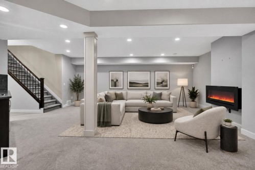 Open-concept recreation area featuring recessed lighting, a wall-mounted electric fireplace, and a carpeted staircase with dark wood railings - 904 Armitage Court Sw, Edmonton, AB 