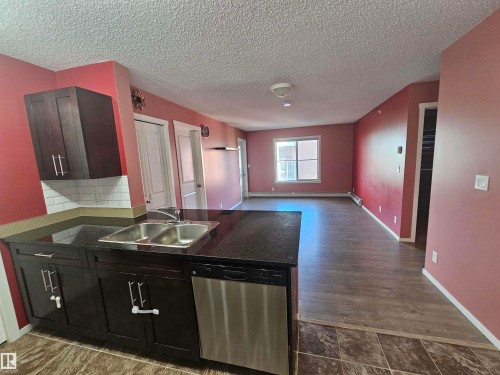 Kitchen featuring dark wood-finish cabinetry, a double basin stainless steel sink, a stainless steel dishwasher, a subway tile backsplash, and dark countertops - #421 5816 Mullen Place Nw, Edmonton, AB 