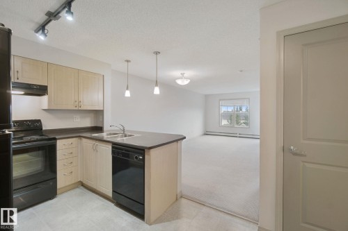Kitchen featuring wood-finish cabinetry, black appliances, a double basin sink, and a peninsula with countertop seating - #406 11449 Ellerslie Rd Sw, Edmonton, AB 