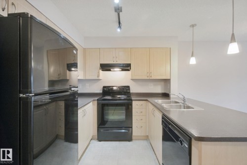 U-shaped kitchen with light wood-finish cabinetry, dark countertops, a double-basin stainless steel sink, and black appliances - #406 11449 Ellerslie Rd Sw, Edmonton, AB 