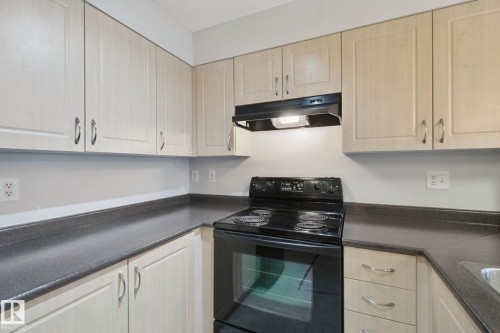 Light wood-finish cabinetry with silver-tone hardware, dark countertops, and a black electric range with an overhead vent - #406 11449 Ellerslie Rd Sw, Edmonton, AB 