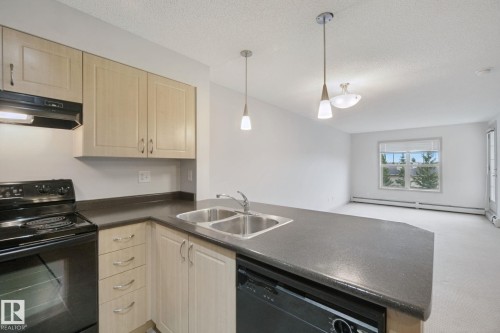 Kitchen featuring light wood-finish cabinetry, dark countertops, and a double basin stainless steel sink - #406 11449 Ellerslie Rd Sw, Edmonton, AB 