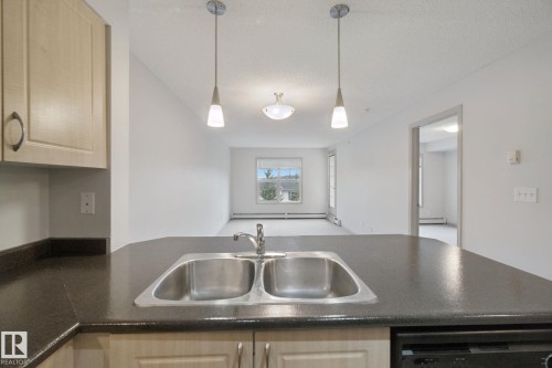 Kitchen island with dual basin stainless steel sink, dark countertop, and pendant lighting - #406 11449 Ellerslie Rd Sw, Edmonton, AB 