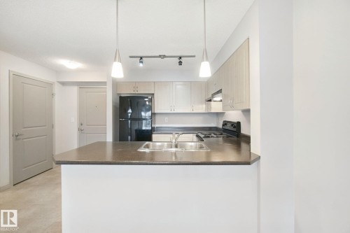 Kitchen featuring light wood-finish cabinetry, dark countertops, a double basin stainless steel sink, and pendant lighting - #406 11449 Ellerslie Rd Sw, Edmonton, AB 