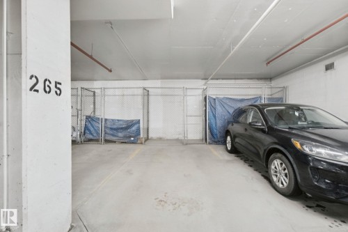 Concrete parking area with a designated space, an adjacent chain-link storage unit, and overhead utility piping - #406 11449 Ellerslie Rd Sw, Edmonton, AB 