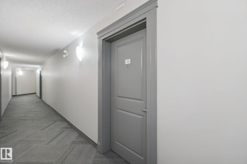 Hallway featuring wall sconces, patterned carpet flooring, and a paneled entry door with substantial trim - #406 11449 Ellerslie Rd Sw, Edmonton, AB 