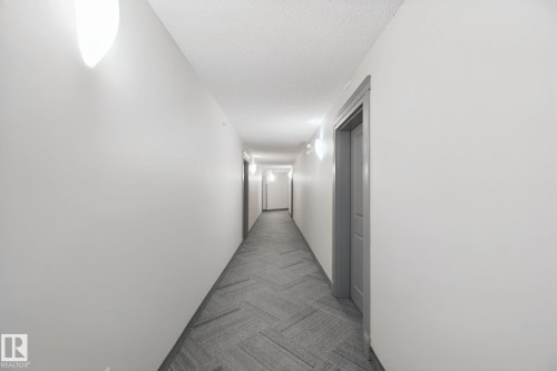 Long hallway with light gray walls, textured ceiling, a patterned carpet runner, and wall-mounted sconces - #406 11449 Ellerslie Rd Sw, Edmonton, AB 