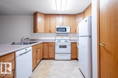 Kitchen featuring light wood cabinetry, white appliances, a double basin stainless steel sink, and light-toned countertops - #203 8912 156 St Nw, Edmonton, AB 