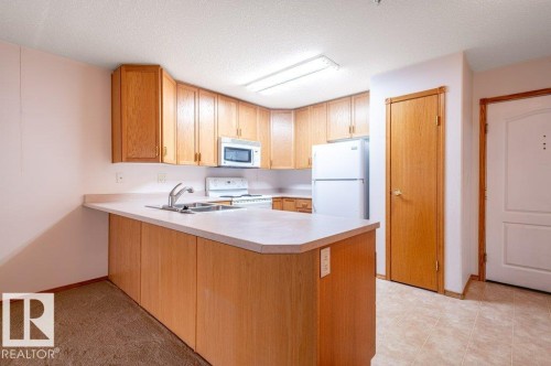 Kitchen featuring wood-finish cabinetry, a double basin sink, a built-in microwave, white appliances, and a tiled flooring area - #203 8912 156 St Nw, Edmonton, AB 