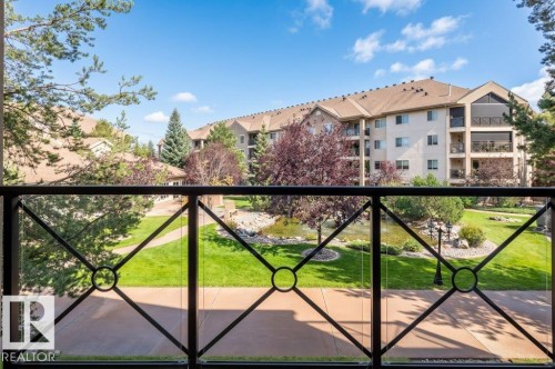 Balcony featuring glass and wrought iron railing, overlooking landscaped grounds with a pond, paved pathways, and mature trees - #203 8912 156 St Nw, Edmonton, AB 