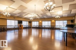 This spacious room featuring wood-finish flooring, multiple chandeliers, acoustic ceiling panels, and extensive window coverage can accommodate many community events like exercise classes, dances, etc. - 