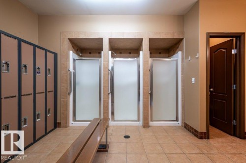 Locker room featuring multiple shower stalls with frosted glass doors, a wood bench, and individual storage lockers - #203 8912 156 St Nw, Edmonton, AB 