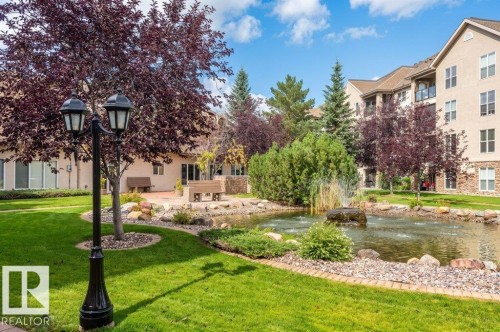 Manicured lawn with a decorative lamppost, a rock-lined pond featuring a fountain, and mature trees - #203 8912 156 St Nw, Edmonton, AB 