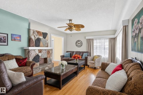 Living room featuring wood-finish flooring, a stone-clad fireplace with a mantel, a ceiling fan, and multiple windows with valances - 12309 80 Street Nw, Edmonton, AB 