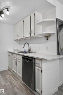 White cabinetry with modern hardware, integrated dishwasher, stainless steel sink with gooseneck faucet, light-toned countertop, and wood-finish flooring - 