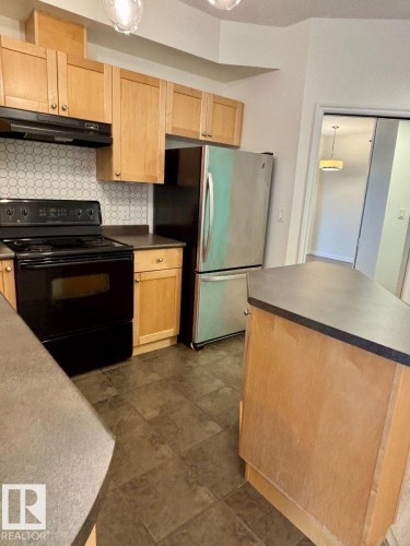 Stainless steel refrigerator, black range, wood-finish cabinetry, honeycomb pattern backsplash, and dark tile flooring - #103 10346 117 St Nw, Edmonton, AB 