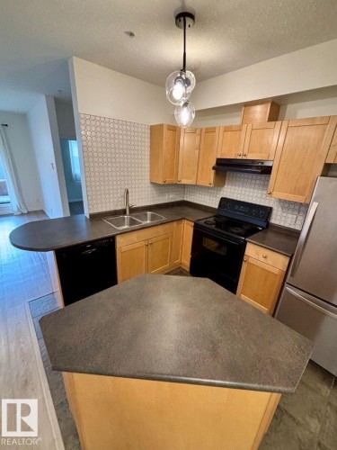 Kitchen featuring light wood-finish cabinetry, dark countertops, a mosaic tile backsplash, and a modern pendant light fixture - #103 10346 117 St Nw, Edmonton, AB 