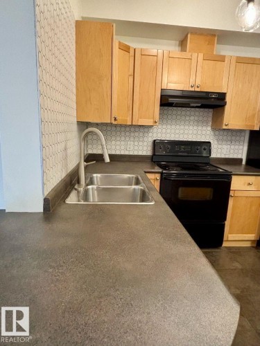 Kitchen featuring a double basin stainless steel sink, light wood cabinetry, matte black range, and hexagonal tile backsplash - #103 10346 117 St Nw, Edmonton, AB 