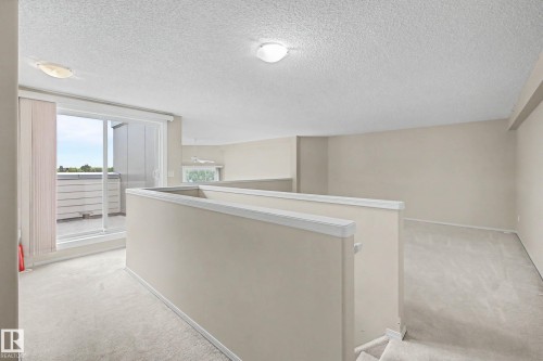 Carpeted interior space with neutral wall tones, featuring a white railing and a sliding glass door providing access to an exterior balcony - #505 7839 96 St Nw, Edmonton, AB 
